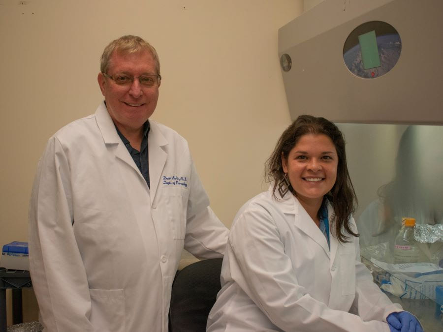 Professor Dean Burkin stands next to Ph.D. student Ariany Oliveira-Santos, who is sitting, in a laboratory room.