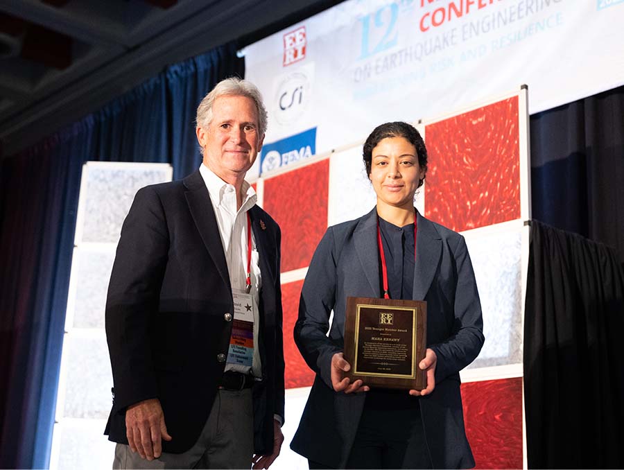 Man and woman in business attire indoors in front of curtain; woman holds an awards plaque.