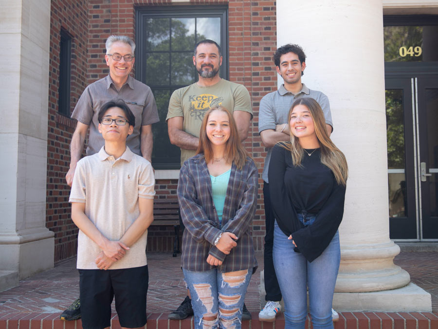 Six people stand in two rows on brick stairs. They are standing in front of a brick building with white columns.