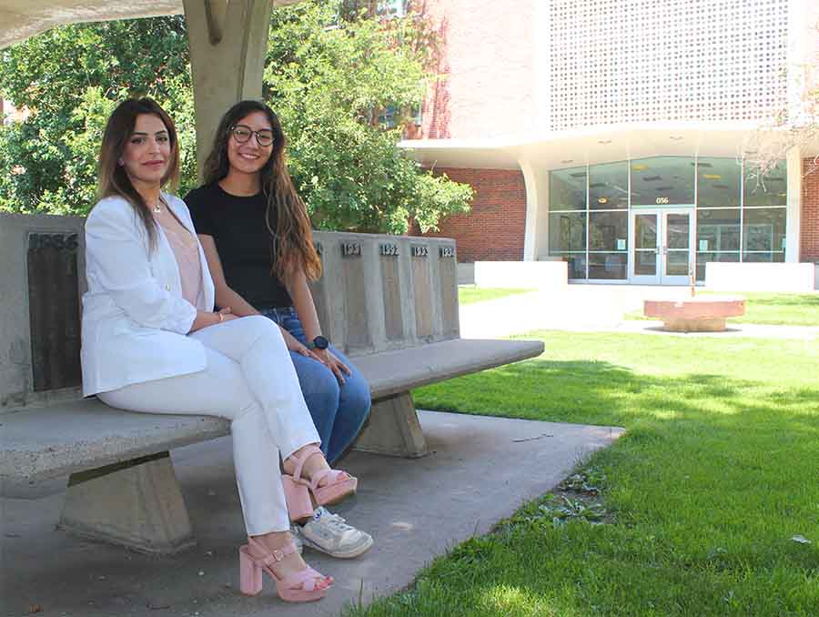 Two women outside sitting on a stone bench in front of a building.