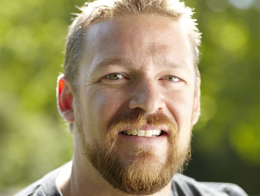 A headshot of Mark Maynard in front of a green nature background.