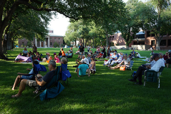 A crowd of people are seated on the University Quad in the shade of the large trees. The brick buildings surrounding the Quad are visible in the background.