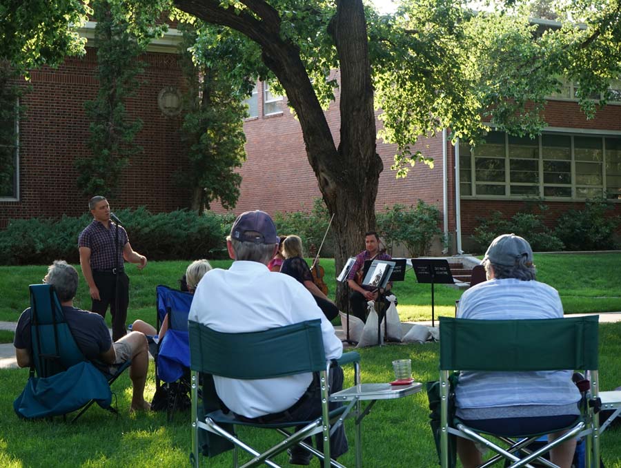A man stands at a microphone in front of a crowd of people sitting in chairs on the University Quad, with several seated people holding instruments beside him. He is standing in front of brick buildings and large trees on a sunny day.