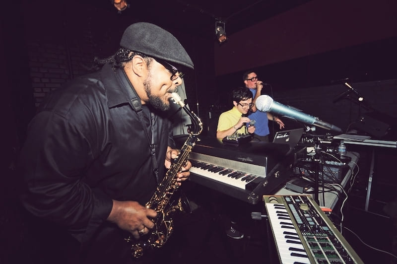 Jamal Tarkington playing the saxophone in front of two pianos and a microphone on stage with two people behind him. 