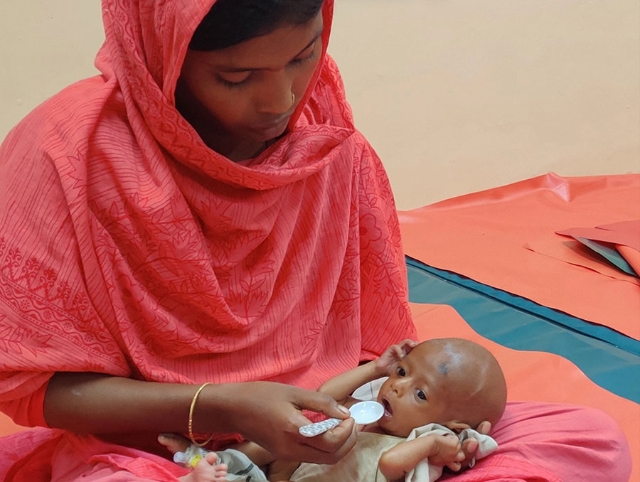 In a hospital, a woman in a bright red shalwar kameez dress and dupattā shawl spoon-feeds an emaciated infant in her lap.