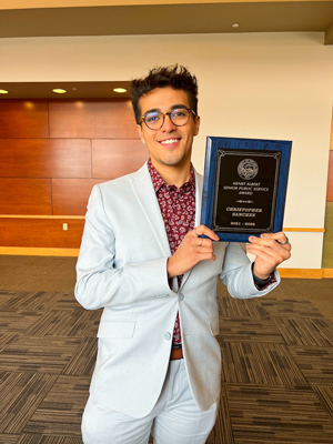 A young man in a light blue suit holds up a plaque with his name on it.