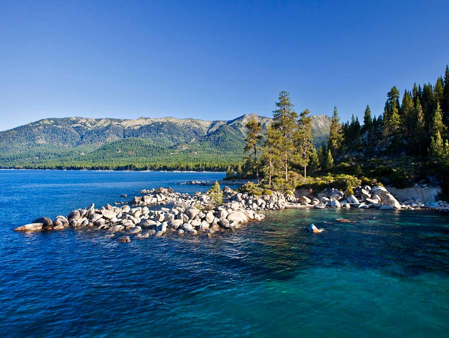 A jetty of rocks spits out to the left with the clear turquoise waters of Lake Tahoe in the foreground and alpine mountains in the background.