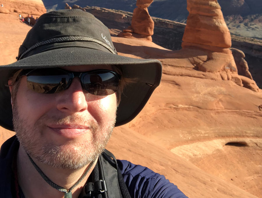 A man wearing a hat and sunglasses stands on red rocks in front of a rock arch.