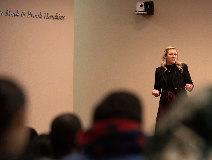 A crowd of people blurred in the foreground watch a presentation by a woman standing on a stage.