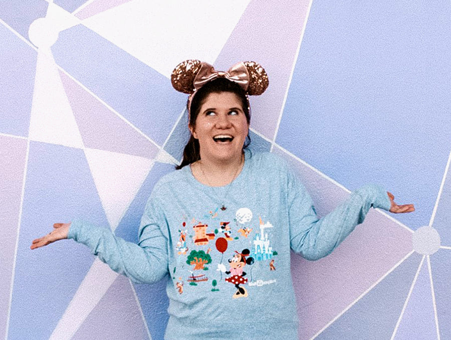 Madeline Potts stands in front of a blue and purple wall wearing pink Disney mouse ears and holding her arms out to the side.