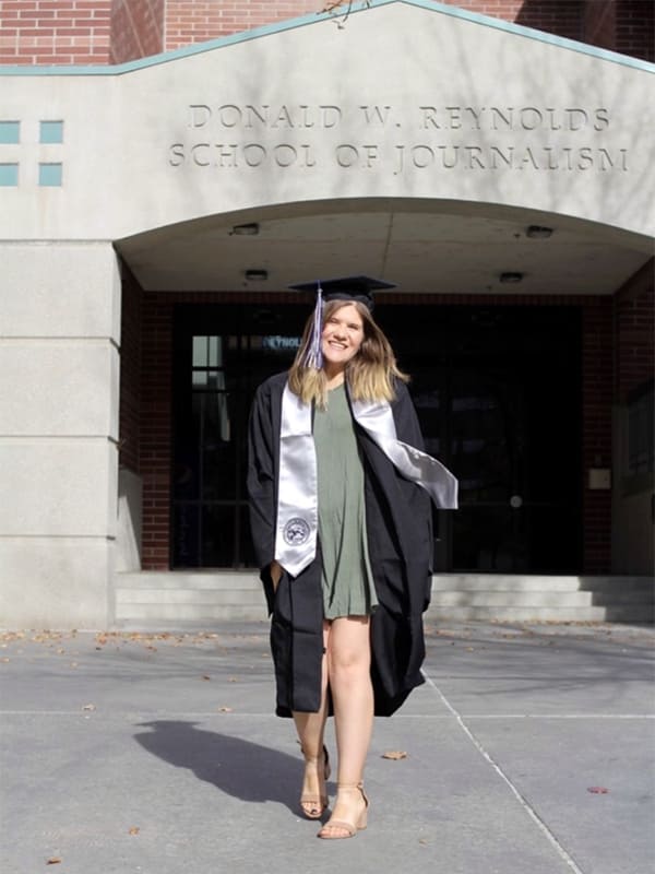 Madeline Potts stands in front of the Reynolds School of Journalism building wearing a graduation robe and cap.