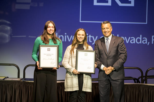 Two young women stand on a stage holding awards next to a man.