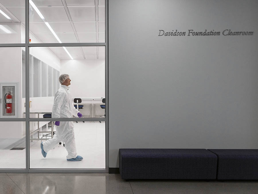 Researcher in scrubs seen through window of Cleanroom lab walking to the right.