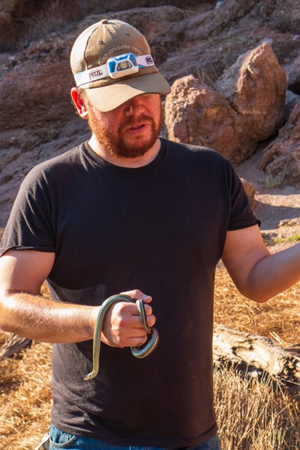 Hallas, wearing a black t-shirt and hat, holds a garter snake.