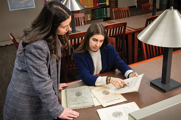 Two women - one leaning over the side of a table and one seated at the table reviewing documents.