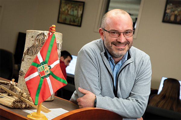 Man leaning on a shelf with arms crossed and a Basque flag to his left