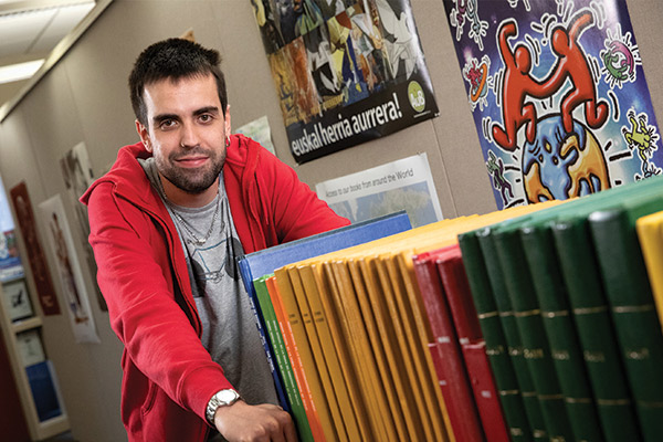 A man wearing a red zip-up hoodie with a grey T-shirt underneath pushes a cart of green, yellow and red books inside of the Jon Bilbao Basque Library.