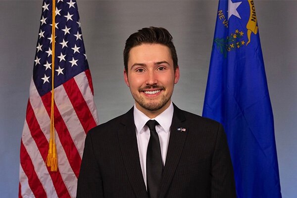 Male college student standing in front of a Nevada flag and American flag wearing a black suit, white shirt and black tie.