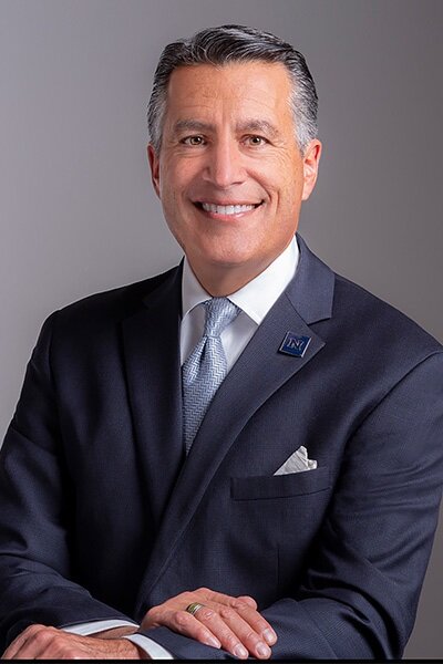 Brian Sandoval portrait. Man wearing a navy blue sport coat, white collared shirt and light blue, patterned tie.