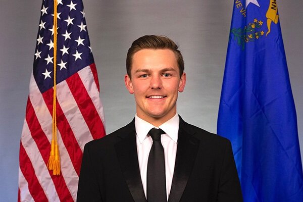 Male college student standing in front of a Nevada flag and American flag wearing a black suit, white shirt and black tie.