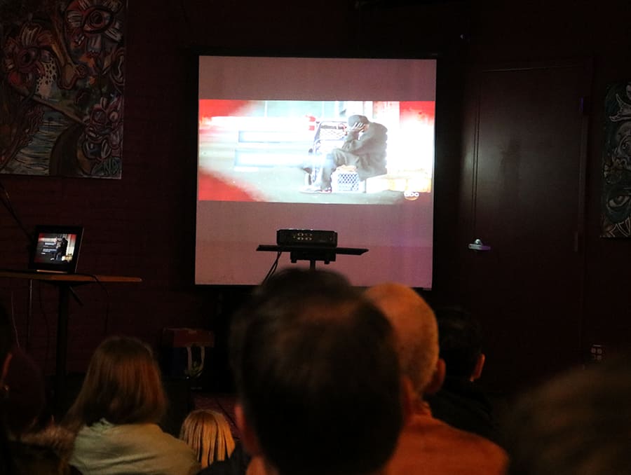 The backs of a few peoples' heads watching a screen that shows a man sitting on a box with his head in his hand.