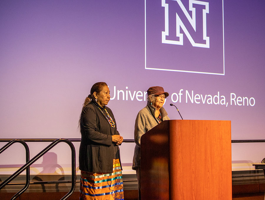 Two individuals speak on stage in front of podium during event