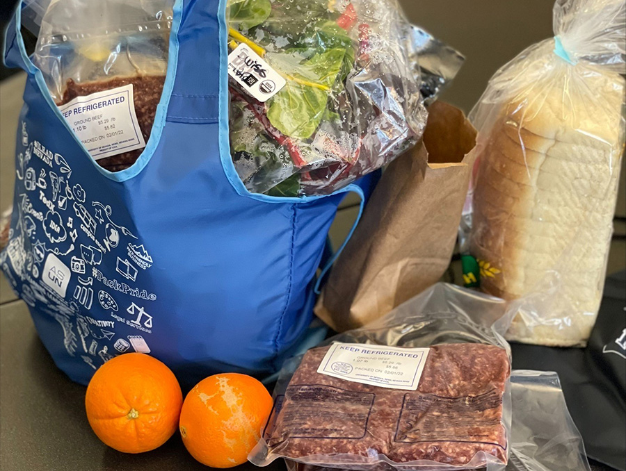 A bag full of fresh ground beef, greens and other food items sits on a table next to a bagged loaf of bread and two clementines and a stack of two packages of ground beef.