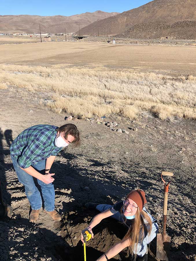 Two people in work boots and jeans use a shovel and other equipment to dig into the soil on farmland. In the background, you can see Veteran's Parkway.