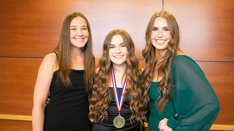 A group of three woman smiling to camera. The woman in the middle wears a medal.