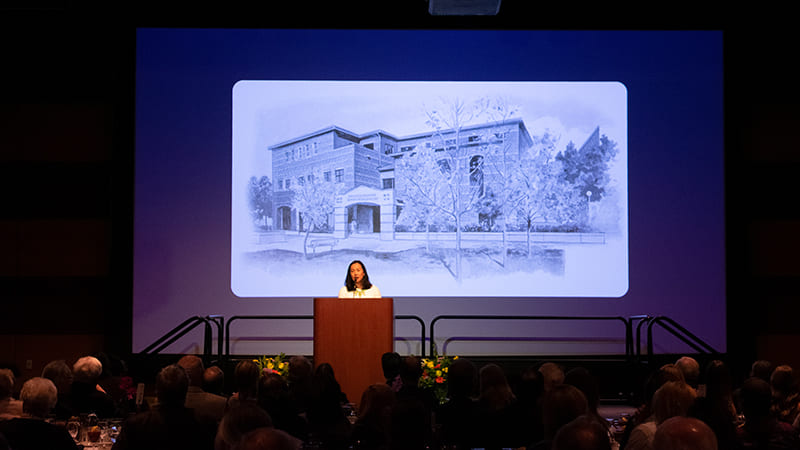 Kristen Go stands on a stage behind a podium in front of a screen that shows the Reynolds School of Journalism building.