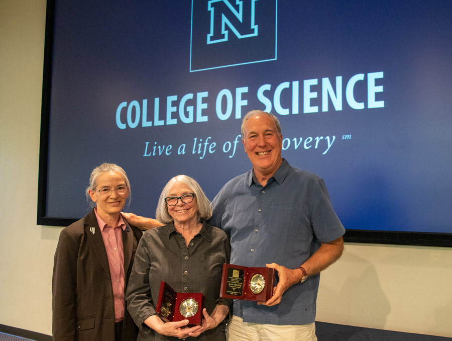 Two women and a man stand, with the woman in the middle and the man holding plaques. They stand in front of a screen that says "College of Science" on the top line and "Live a life of discovery" on the bottom line in italics. The Nevada Block N is above the words.