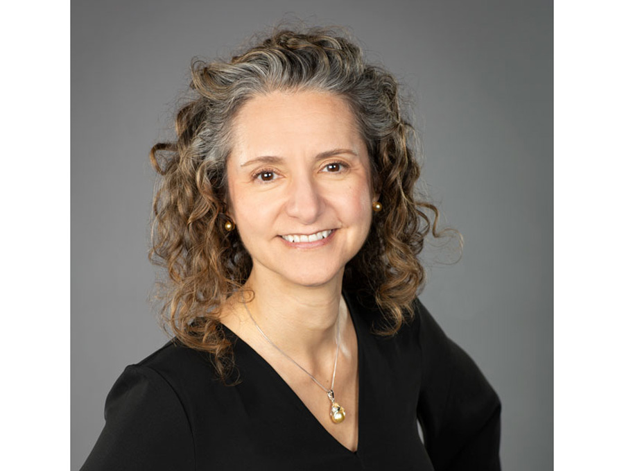 de Bettencourt-Dias wears a black shirt with a necklace and smiles in front of a gray background.