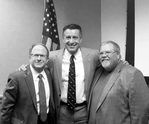 A black and white photo of Alan Stavitsky, Brian Sandoval and Guy Clifton standing together and smiling to camera.