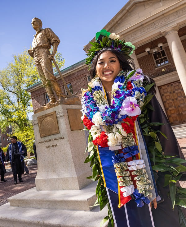 A graduate with many flowers draped around their neck with a freshly restored Mackay Statue in the background