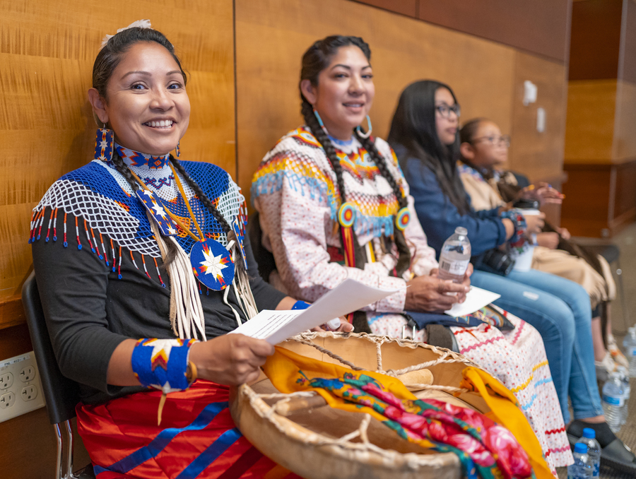 Four women dressed in traditional Native American outfits sit in a ballroom holding instruments smiling at the camera..