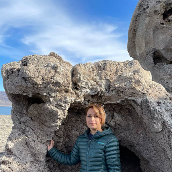 A woman stands in front of a large tufa structure, wearing a puffy teal coat.