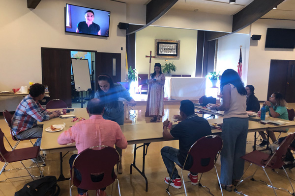 A group of people stand and sit and eat at tables. Another person is on a television screen. At the front of the room is a church altar and American flag.