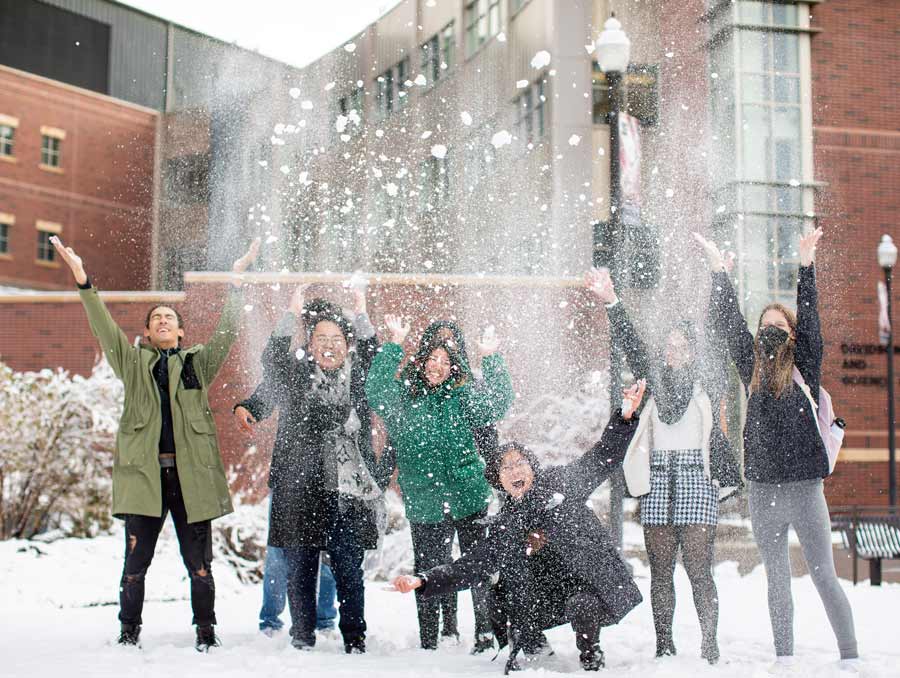 Eight students bundled up for winter weather raise their arms up and throw snow in the air in front of the Davidson Mathematics and Science Center.