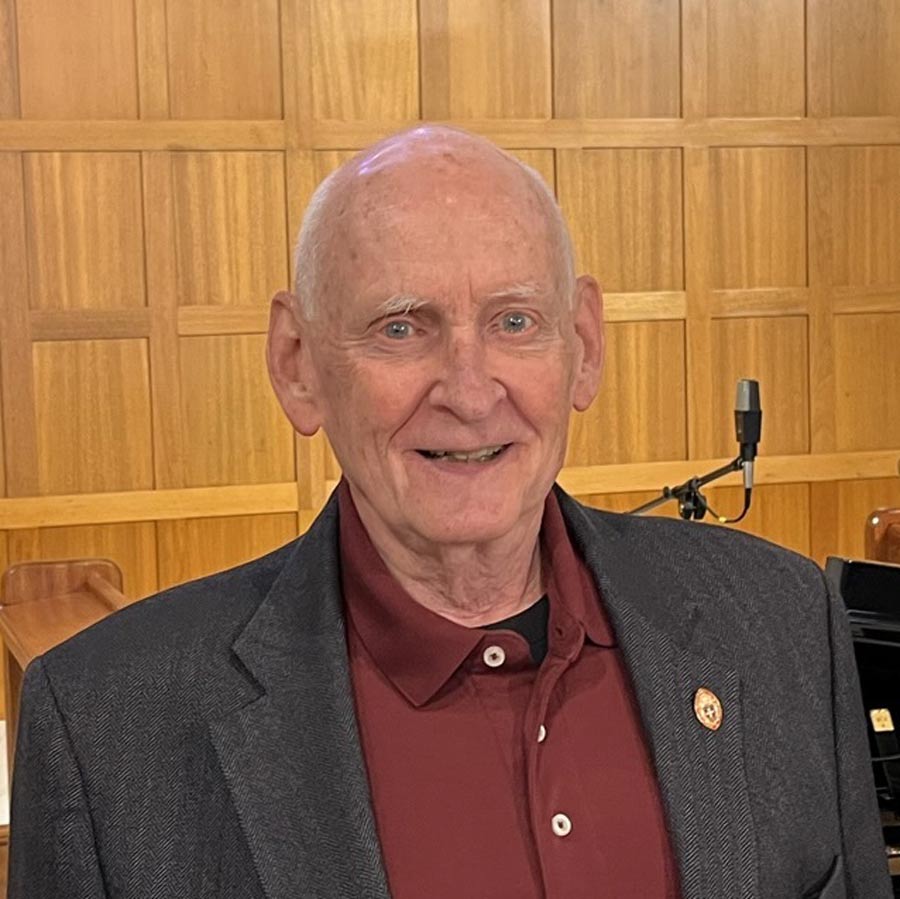 A man wearing a grey jacket over a maroon polo shirt smiles in front of wooden paneling and instruments.