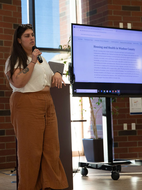 A woman wearing rust-colored pants and a white shirt stands near a podium and screen speaking into a microphone. The header text on the screen reads, "Housing and Health in Washoe County."