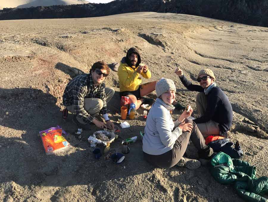 Four people sit on the ground surrounded by jackets and cooking items. They are all facing the camera and smiling, dressed warmly.