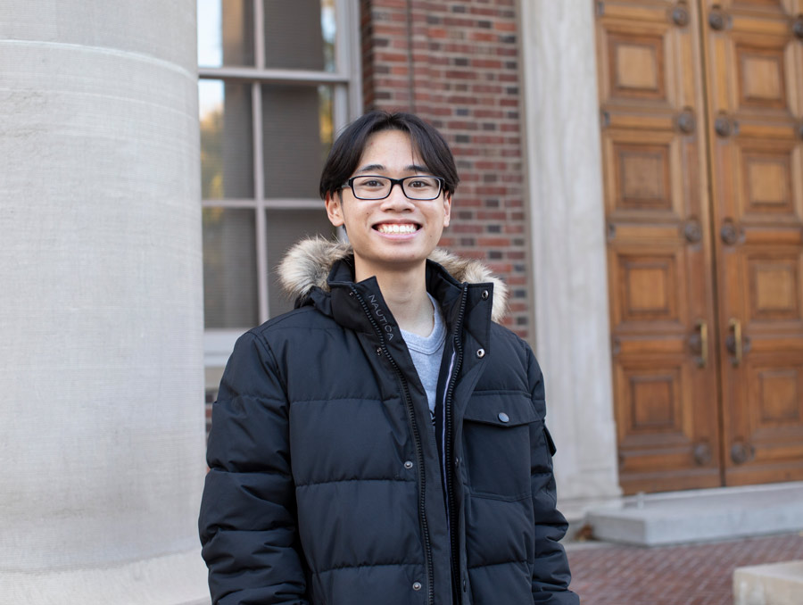 Jonathan Taasan smiles, wearing glasses and a black coat and standing in front of the Mackay School of Mines building.