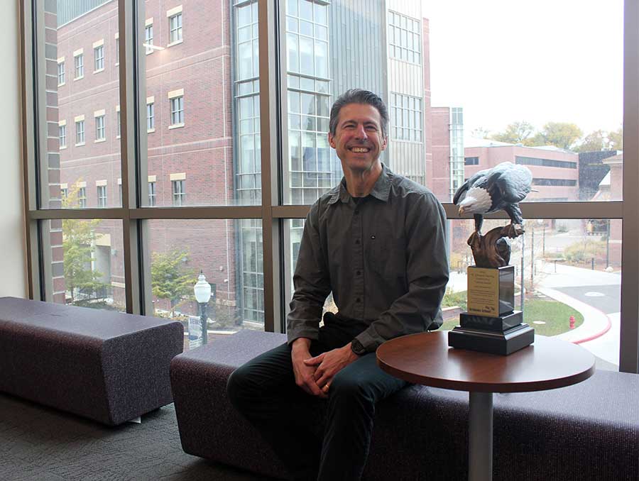 Professor Miles Greiner seated indoors, next to the Greiner award trophy, with a window behind him.