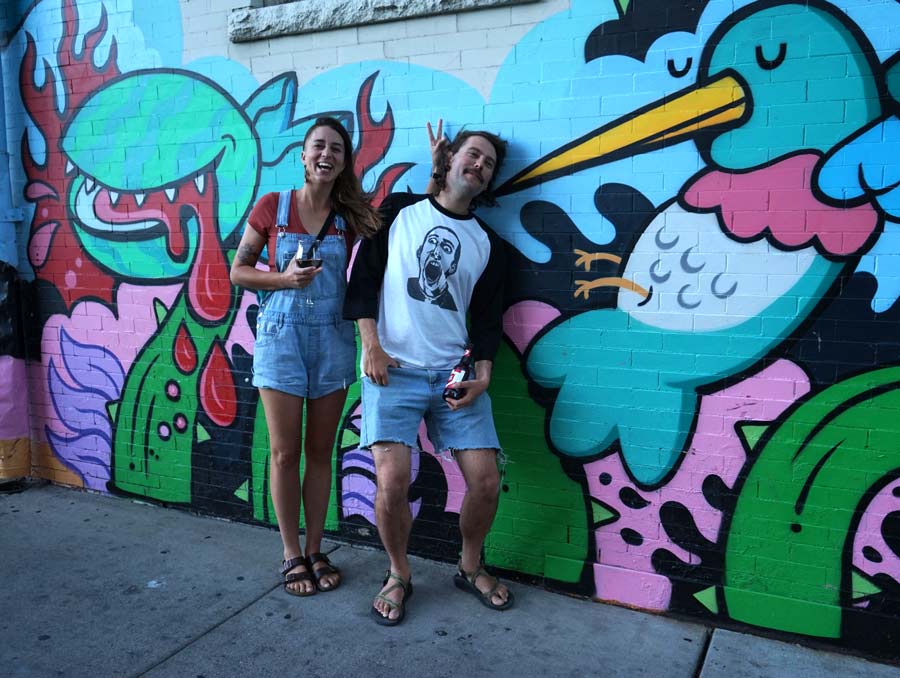 A woman and man lean against a brightly-colored mural. The woman is laughing, holding up two fingers behind the man's head, while he leans in an awkward position into the mural.