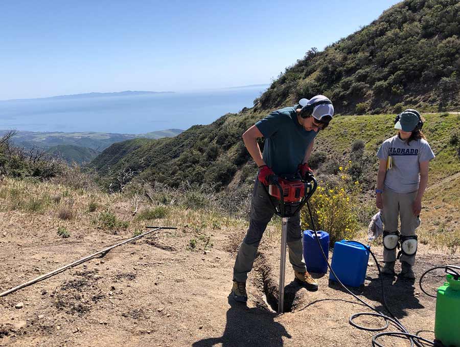 A person stands in a clearing holding a large drill. Another person stands nearby with a notebook under their arm. They are standing in a mountainous area with the ocean visible in the background.