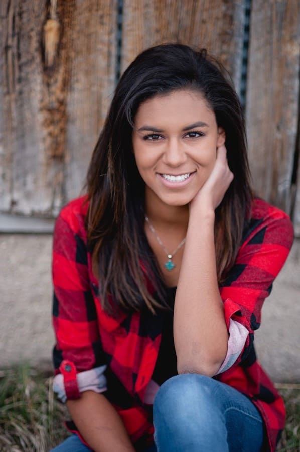 Young woman in red-flannel patterned shirt and blue wash jeans sitting in the grass in front of a brown fence.