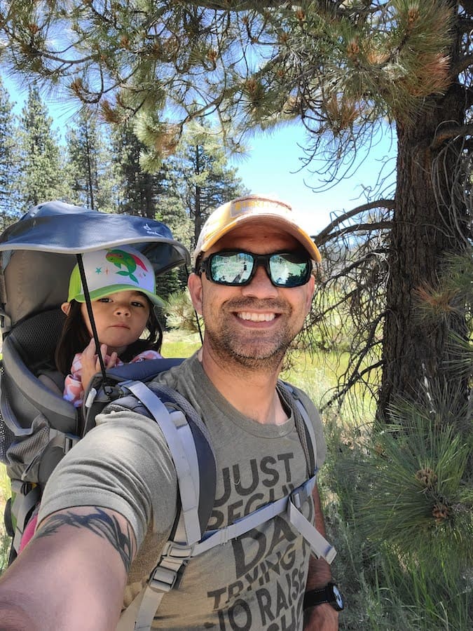 Man and his toddler-aged child on his back, inside a backpack-type sling, on a hike with trees in the background.