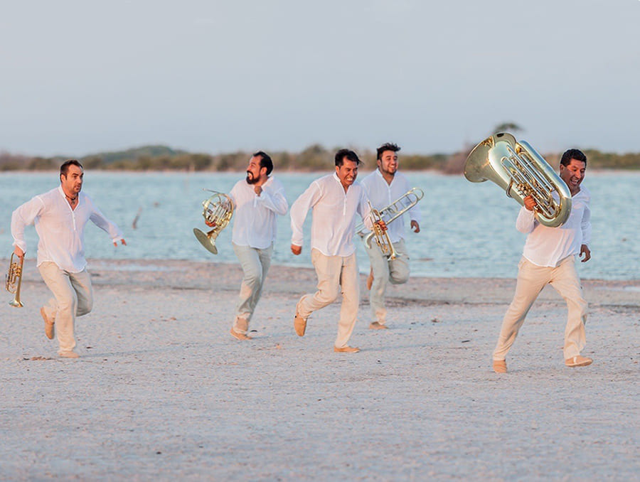 Members of M5 Mexican Brass running on the beach carrying their instruments.