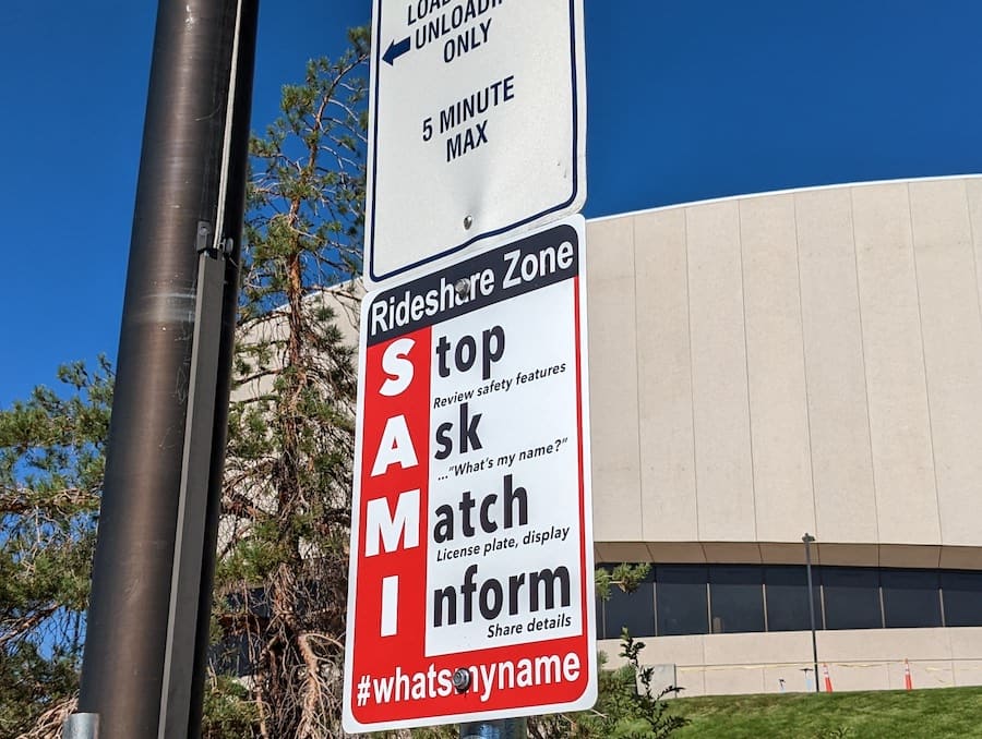 Red, black and white sign (with acronym S.A.M.I.) posted on pole in front of brown building with blue sky in the background.