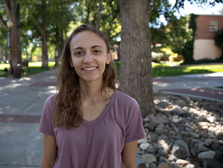 A woman wearing a mauve shirt smiles, with brick buildings and large trees over green grass in the background.